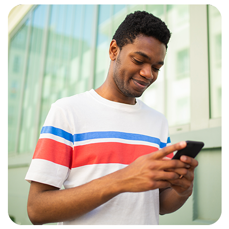 Jovem negro, sorrindo, em pé na rua olhando o celular.